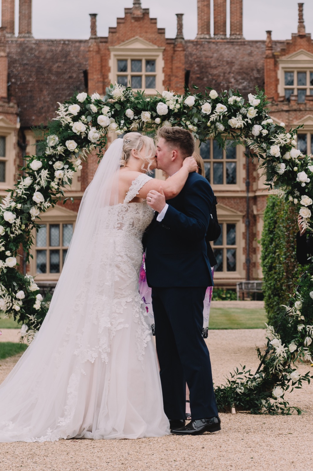 Couple kissing beneath a moon gate arch of peonies, roses and greenery at Haughley Park Barn